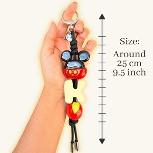 May include: A Mickey Mouse-themed keychain, approximately 9.5 inches long. The keychain features a black and red Mickey Mouse head, the letters "M-I-C-K-E-Y" in cream, and a red and yellow design. It includes a silver keyring and black cord.