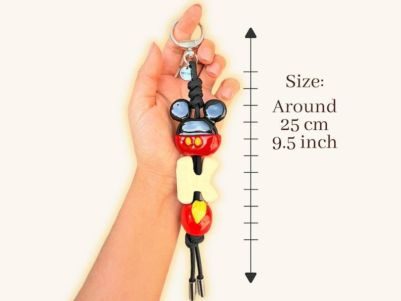 May include: A Mickey Mouse-themed keychain, approximately 9.5 inches long. The keychain features a black and red Mickey Mouse head, the letters "M-I-C-K-E-Y" in cream, and a red and yellow design. It includes a silver keyring and black cord.