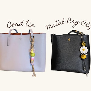 May include: Two handbags: a light gray tote with a cord tie and a black satchel with a metal bag clip. The cord tie has colorful beads and a tassel. The metal bag clip features a daisy and a smiling bee.