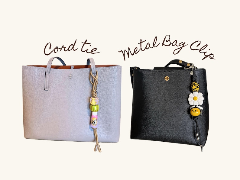 May include: Two handbags: a light gray tote with a cord tie and a black satchel with a metal bag clip. The cord tie has colorful beads and a tassel. The metal bag clip features a daisy and a smiling bee.