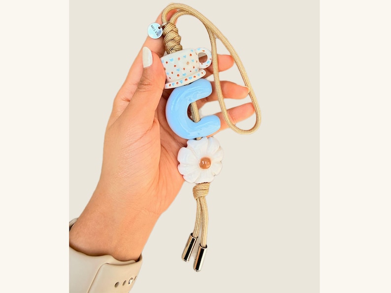 May include: A beige cord phone charm with a blue letter "C", a white daisy, and a small teacup charm. The charm has gold-colored metal accents and a small tag.