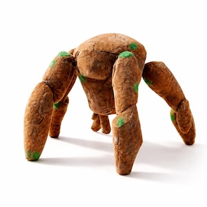 Puede incluir: Un juguete de cangrejo marrón con seis patas articuladas y un cuerpo redondeado. El cangrejo tiene detalles verdes en las articulaciones y está sobre un fondo blanco. El juguete está diseñado para parecerse a un crustáceo.
