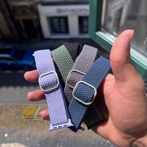 Puede incluir: Un conjunto de cuatro correas de reloj elásticas trenzadas en lavanda, verde oliva, gris carbón y azul marino. Cada correa cuenta con una hebilla deslizante rectangular de metal plateado y una pieza de conexión a juego para un reloj inteligente.