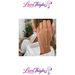 Pode incluir: Uma corrente de mão dourada composta por uma corrente delicada que liga um anel a uma pulseira. A peça de joalharia é adornada com três pequenas pedras transparentes de corte marquise, espaçadas uniformemente ao longo da corrente.