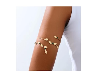 May include: A gold-toned arm cuff featuring a delicate vine design with multiple metallic leaves and small, round, dark green accents. The jewelry wraps around the upper arm in a spiral shape, showcasing a polished finish and a slender, flexible band structure.