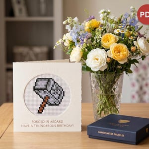 Può includere: Biglietto di auguri color crema con un disegno a punto croce del martello di Thor, Mjolnir. Il testo del biglietto recita: "Forgiato ad Asgard, Buon Compleanno Tuonante!" Un vaso di rose gialle e bianche è vicino.