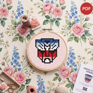 May include: A cross-stitch project featuring a red, white, and blue geometric design within a wooden embroidery hoop. The design is set against a floral background with pink roses and blue flowers. The image includes spools of thread and a booklet titled "Cross-Stitch Heroes".