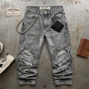 Könnte beinhalten: Graue Jeans aus Denim mit einem Design aus geschichteten Paneelen mit ausgefransten Kanten, einer silbernen Metallkette an der Gürtelschlaufe und einer schwarzen Geldbörse. Das Etikett am Bund zeigt FORGED DENIM. Dazu graue und weiße Sneaker, ein braunes Notizbuch und ein Stift.