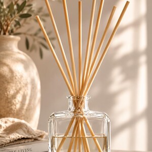 May include: A clear, square glass reed diffuser bottle filled with a light-colored liquid. Several thin, light-tan wooden reeds are inserted into the bottle, fanning outward from the narrow neck. The glass container has a smooth, transparent finish.