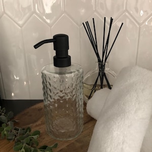 May include: A clear glass soap dispenser with a black pump sits on a wooden surface. Beside it is a clear glass diffuser with black reeds and a rolled white towel. The background features white, geometric tiles.