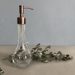 May include: A clear glass soap dispenser with a teardrop-shaped base and a metallic copper-colored pump mechanism. The dispenser features a long, slender neck and a wide, rounded bottom, providing a functional and sleek design for liquid soap.