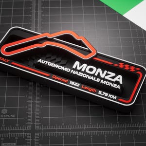 May include: A black rectangular 3D wall plaque featuring the Monza race track layout in orange and white. The text reads ITALY, MONZA, AUTODROMO NAZIONALE MONZA, Opened 1922, and Length 5.79 KM, with a checkered flag graphic on the right side.