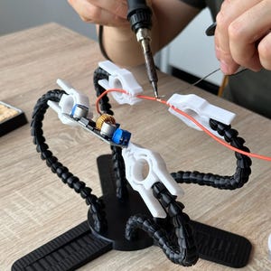 Peut inclure: Station de soudure noire et blanche avec des bras flexibles et des pinces blanches. Une personne utilise un fer à souder pour connecter des fils. La station maintient des composants électroniques.