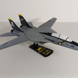 Może przedstawiać: Szary model samolotu F-14 Tomcat z czarnymi i żółtymi akcentami. Model ma numer 103 i emblemat czaszki i skrzyżowanych piszczeli. Stoi na czarnym stojaku z napisem "ApexCraft F-14 TOMCAT".