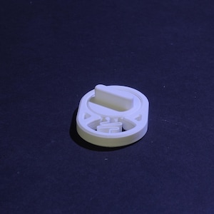 May include: A white, circular, 3D-printed plastic component featuring a central raised tab and a lower section with an open, geometric cutout design. The object has a smooth, matte finish and a flat base, showing a distinct structural form.