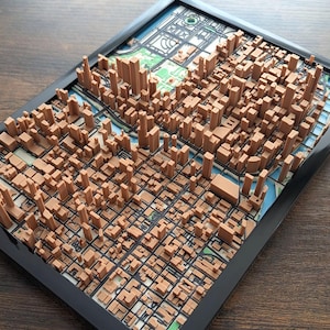 May include: A framed 3D city map showing a detailed brown model of buildings and infrastructure. The map is set against a backdrop of a colorful street map, all encased in a black frame. The artwork provides a unique perspective of an urban landscape.
