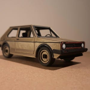 Op de afbeelding: Een gedetailleerd modelauto in goud en zwart. Het miniatuurvoertuig heeft een getextureerde carrosserie, zwarte banden en een rode accent op de grille. De auto is een klassiek hatchback-ontwerp.