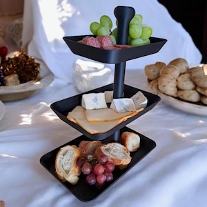 May include: A three-tiered black serving tray laden with food. The top tier holds green grapes and sliced meat. The middle tier has cheese and sliced meat. The bottom tier has bread and red grapes. The tray is on a white tablecloth.