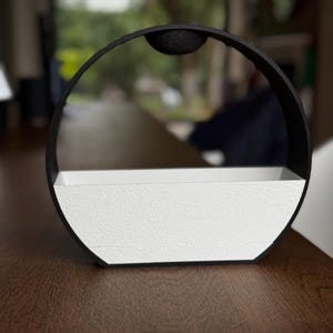 Peut inclure: Un pot de fleurs moderne à deux tons, avec un cadre circulaire noir et un récipient rectangulaire blanc. Le pot a une surface texturée et un petit accent noir rond en haut. Le design est minimaliste et convient aux petites plantes ou objets décoratifs.