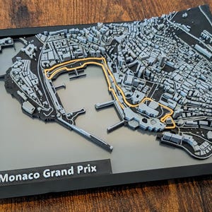 Peut inclure: Une carte imprimée en 3D du circuit du Grand Prix de Monaco, avec une représentation détaillée de la ville et du tracé. La piste est mise en évidence en or, encadrée de noir. Le texte "Monaco Grand Prix" est imprimé en bas.