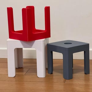 Könnte beinhalten: Drei kleine, quadratische Hocker in Rot, Weiß und Grau. Der rote Hocker ist auf dem weißen gestapelt, und der graue Hocker hat ein Loch in der Mitte. Alle haben ein minimalistisches Design mit vier Beinen.