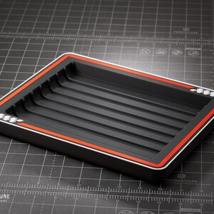 Peut inclure: Un plateau rectangulaire noir avec un intérieur strié. Le plateau a une bordure rouge et blanche. Le logo "FORMSTUBE 3D DESIGN & PRINT" est visible dans le coin inférieur gauche. Le plateau est sur une surface grise.