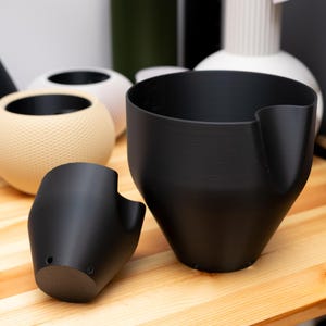 Può includere: Una collezione di vasi moderni e minimalisti in varie forme e colori. In primo piano ci sono due vasi neri, uno con un'estensione simile a una maniglia. Altri vasi includono una sfera beige e un design bianco a coste. Gli oggetti sono esposti su una superficie di legno.