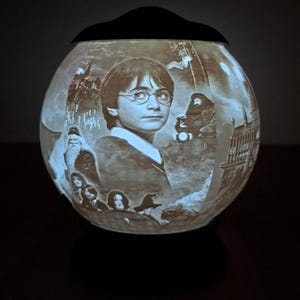 May include: A spherical decorative jar with a black lid, illuminated from within. The jar's surface is etched with scenes and characters from Harry Potter, including Harry himself. The jar emits a soft, warm light, creating a magical effect.