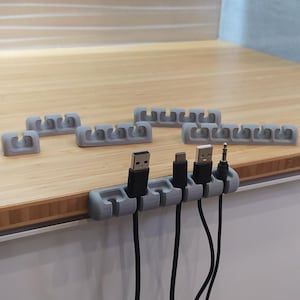 Peut inclure: Plusieurs organiseurs de câbles gris sont présentés sur une surface en bois. Chaque organisateur comporte plusieurs fentes pour maintenir divers câbles, y compris des câbles USB et audio. Les organisateurs sont conçus pour ranger les câbles.