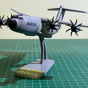 Peut inclure: Maquette grise d'un avion A400M Atlas avec quatre hélices noires et des pointes jaunes. Le modèle est monté sur un socle gris avec le texte "A400M Atlas". L'avion présente un design détaillé, sur fond de grille verte.