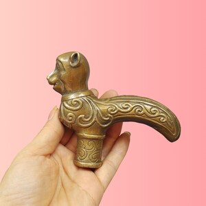 May include: A brass cane handle shaped like a stylized animal head with a curved neck and body. The metal surface features intricate, embossed scrollwork patterns across the neck and the cylindrical base, showcasing a warm, metallic gold tone.