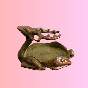 May include: A bronze-toned metal dish shaped like a resting deer with its head turned back and antlers extending over the center. The bowl area is circular and shallow, featuring a smooth, weathered metallic surface with dark brown patina accents.