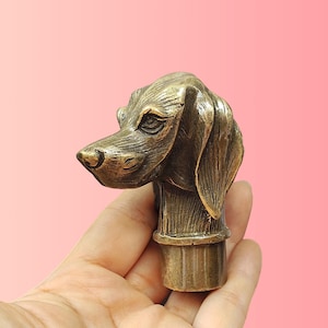 Könnte beinhalten: Eine metallene, bronzefarbene Hundekopfskulptur mit detaillierten Gesichtszügen, hängenden Ohren und einer strukturierten Schnauze. Die Skulptur hat einen zylindrischen Sockel und ist mit einem verwitterten, antiken Finish versehen.