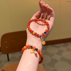 Könnte beinhalten: Zwei Armbänder mit orangefarbenen und gelben Perlen, akzentuiert mit kleinen blauen und gelben Anhängern. Die Armbänder werden am Handgelenk getragen und zeigen das Design und die Farbe des Schmucks.
