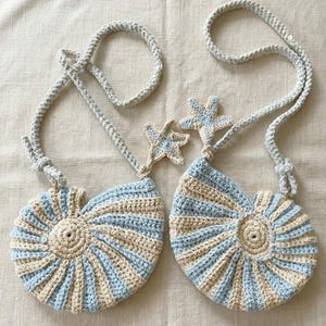 Puede incluir: Dos bolsos hechos a mano de ganchillo con forma de concha marina, a rayas azul claro y crema. Cada bolso tiene una correa azul claro a juego y un adorno de estrella de mar. Los bolsos están diseñados para llevarse al hombro.