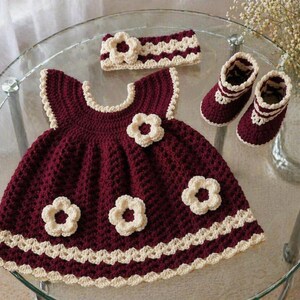 May include: A handmade crochet baby outfit in burgundy and cream. The set includes a dress with flower appliqu&eacute;s, a matching headband, and booties. The dress and booties have a scalloped cream trim. The outfit is displayed on a glass table.