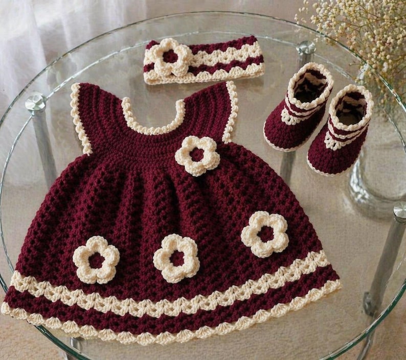 May include: A handmade crochet baby outfit in burgundy and cream. The set includes a dress with flower appliqu&eacute;s, a matching headband, and booties. The dress and booties have a scalloped cream trim. The outfit is displayed on a glass table.