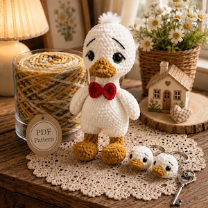 May include: A white crocheted duck doll with a yellow beak and feet, wearing a red bow tie, accompanied by two small duck head keychains. A round tag labelled PDF Pattern is attached to a glass jar filled with variegated yellow and white yarn.