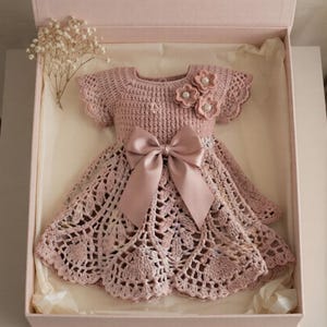 May include: A dusty pink crochet dress featuring a textured bodice, short sleeves, and a flared skirt with an intricate openwork pattern. The dress is adorned with three small crochet flowers with pearl centers and a large satin ribbon bow at the waist.
