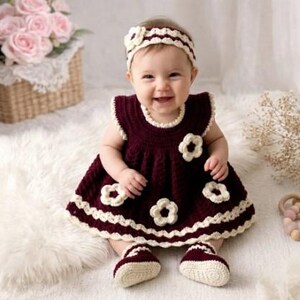 May include: A baby wearing a burgundy crochet dress with white flower accents, matching booties, and a headband. The dress has a scalloped hem and neckline. The baby is sitting on a white fluffy surface.