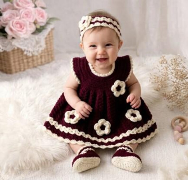 May include: A baby wearing a burgundy crochet dress with white flower accents, matching booties, and a headband. The dress has a scalloped hem and neckline. The baby is sitting on a white fluffy surface.