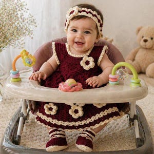 May include: A baby in a burgundy crochet dress with cream-colored trim and flower accents, matching headband and booties. The baby is seated in a white and gray baby walker with colorful toys. A teddy bear sits in the background.
