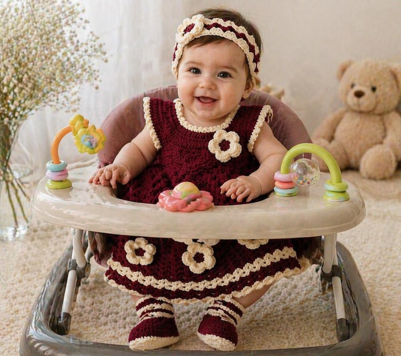 May include: A baby in a burgundy crochet dress with cream-colored trim and flower accents, matching headband and booties. The baby is seated in a white and gray baby walker with colorful toys. A teddy bear sits in the background.