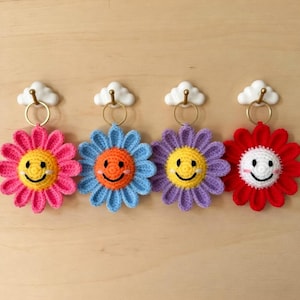 May include: Four crochet flower keyrings in pink, blue, purple, and red, each with a smiling face centre. They hang from white cloud-shaped hooks with gold-coloured rings. The background is a light wood surface.