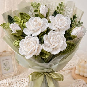 Op de afbeelding: Een boeket handgemaakte gehaakte bloemen, met grote witte rozen en groene bladeren. De bloemen zijn verpakt in lichtgroen papier en vastgebonden met een bijpassend lint. Een uniek, handgemaakt cadeau.