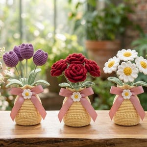 May include: Three crochet flower arrangements in yellow pots with pink ribbons. The arrangements feature purple tulips, red roses, and white daisies with yellow centers. The flowers are displayed on a wooden surface.