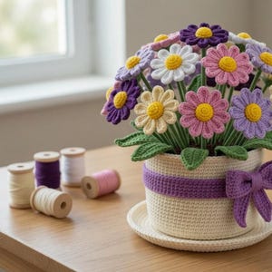 May include: A handcrafted crochet flower arrangement in a cream-colored pot with a purple ribbon and bow. The arrangement features colorful daisy-like flowers in shades of purple, pink, white, and yellow. Green leaves are also visible. Spools of thread and crochet hooks are on the table.