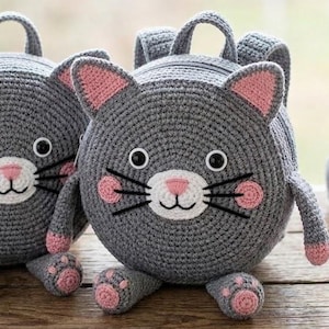 May include: Two gray crochet cat-shaped backpacks with pink accents on the ears, cheeks, and paws. Each backpack has a handle and adjustable straps. The cat faces have black whiskers and white and pink noses. A ball of gray yarn and a pink crochet hook are also visible.