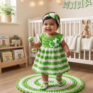 Könnte beinhalten: Ein gehäkeltes Babykleid mit horizontalen grünen und weißen Streifen, einem gewellten Saum und einer großen Blumenapplikation auf der Brust. Das Set enthält ein passendes weißes Stirnband mit Blumen und gehäkelte Schuhe mit Blumenverzierungen.
