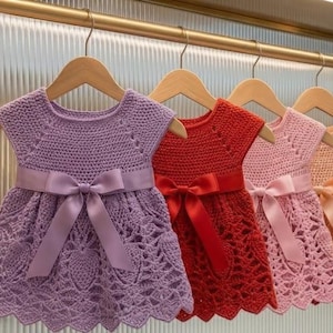 May include: A row of five sleeveless crochet dresses hanging on wooden hangers. Each dress features a distinct solid colour including lavender, red, light pink, peach, and light blue, with a matching satin ribbon bow tied at the waist.